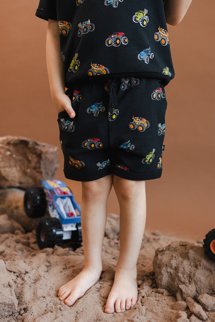 Boy wearing Black Jogger shorts in Monster Truck print on sand and rocks with toy trucks