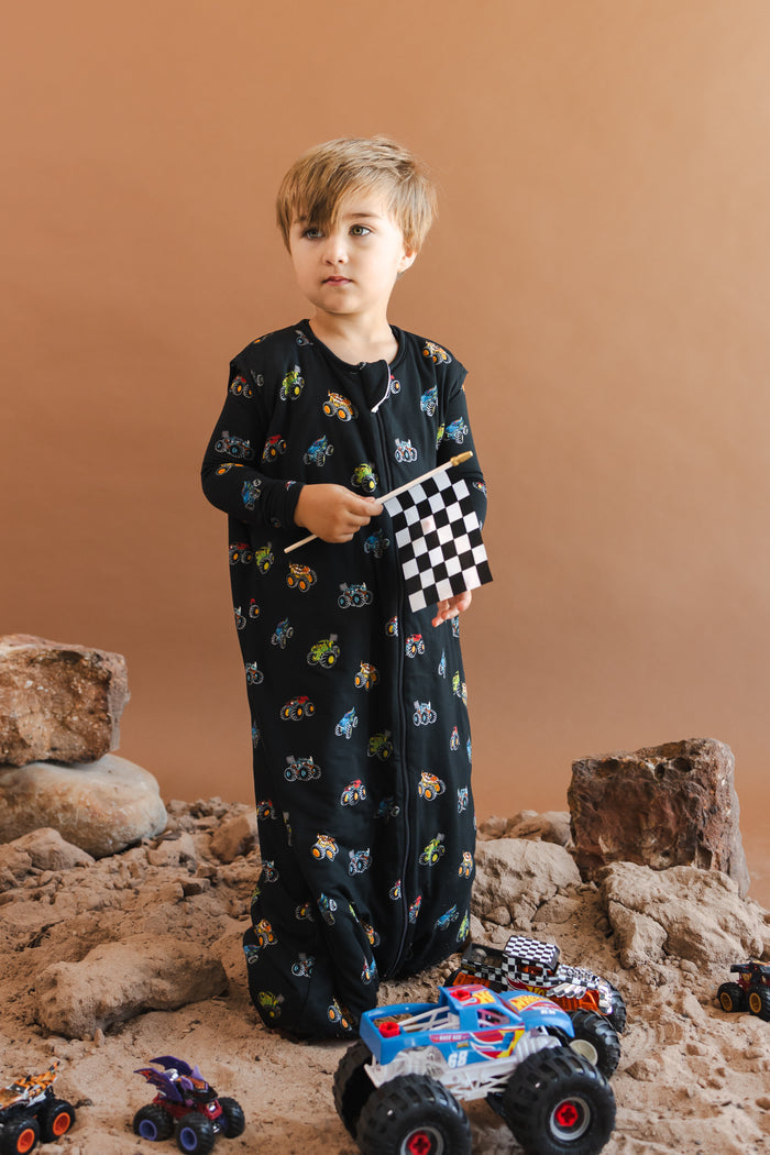 Boy wearing black Sleep Bag Walker with Monster Truck print standing on sand and rocks