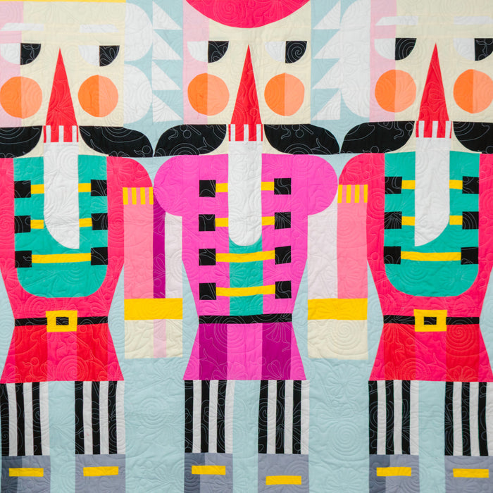 A colorful quilted wall hanging or blanket featuring three stylized nutcracker figures in bright pink, red, teal, black, white, and yellow on a pale blue background, shown hanging indoors against a dark gray wall with a small tree and red ladder visible at the edges.
