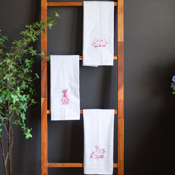White towels with red embroidery on a wooden ladder against a dark wall with plants and flowers.