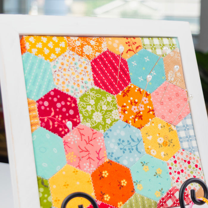 A white-framed hexagonal patchwork quilt art print displayed on a black metal stand in a home setting with a blurred background of plants and books.