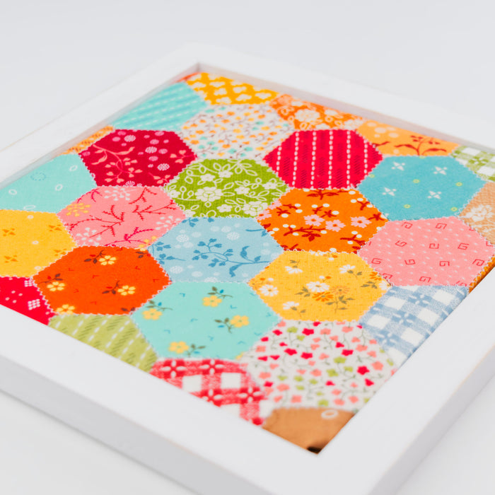 A square white framed art piece featuring a colorful patchwork quilt design made of hexagonal fabric blocks in various patterns and colors, photographed on a clean white background.