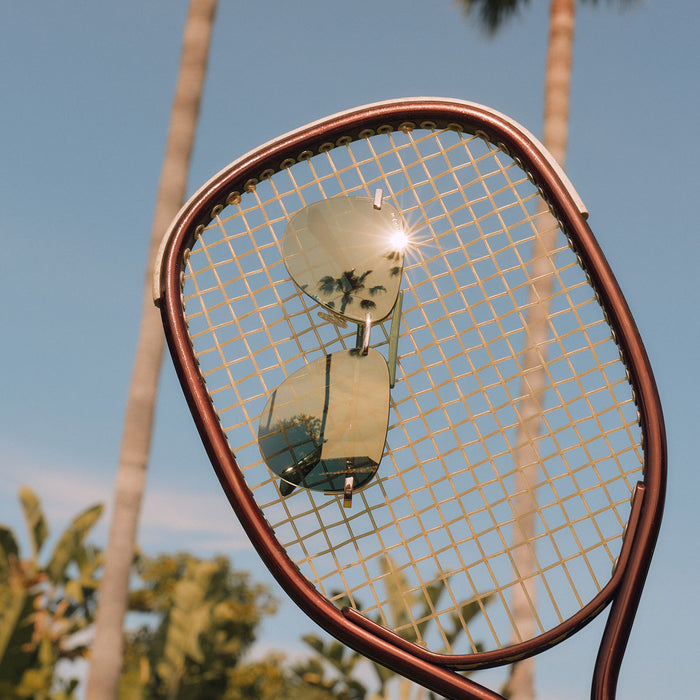 still life - tahoe gold aloe green aviator sunglasses held up on a tennis racket reflecting the view