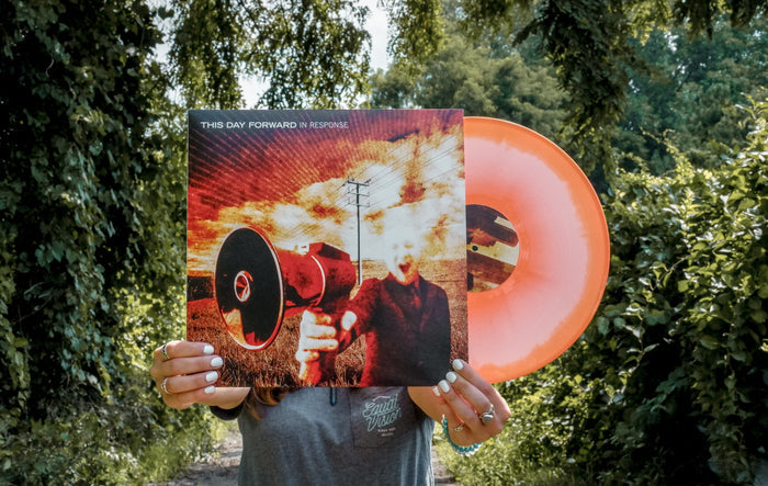 A person stands on a leafy path, holding a limited edition orange/white Therefore I Am vinyl with transparent center and its album cover—In Response—in front of their face so the art aligns with their head.