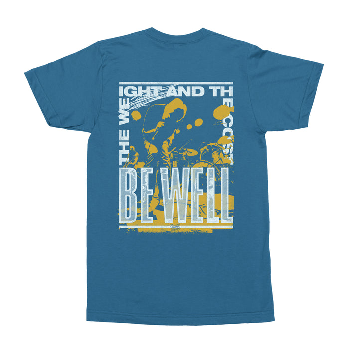 Blue t shirt against white background. Back of t shirt has words printed in white making a rectangle that say "THE WEIGHT AND THE COST" at the top and "BE WELL" at the bottom. Behind the words is a silhouette picture of an individual performing on stage in yellow.
