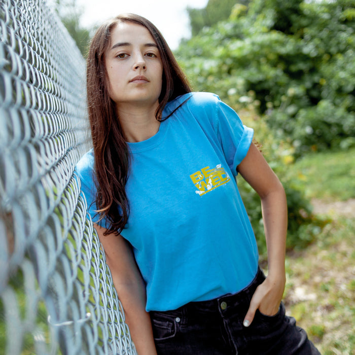 Blue t shirt with "BE WELL" printed in bold yellow font in upper right corner. White shapes are placed behind the words. Individual is modeling t shirt and is leaning against a fence outside with trees in the background.