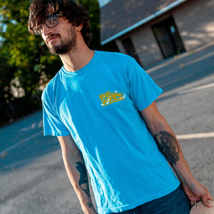 Blue t shirt with "BE WELL" printed in bold yellow font in upper right corner. White shapes are placed behind the words. Individual with tattoos is modeling t shirt outside with a building in the background.