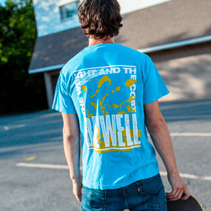 Individual holding a skateboard is modeling blue t shirt outside with a building in the background. Back of t shirt has words printed in white making a rectangle that say "THE WEIGHT AND THE COST" at the top and "BE WELL" at the bottom. Behind the words is a silhouette picture of an individual performing on stage in yellow.