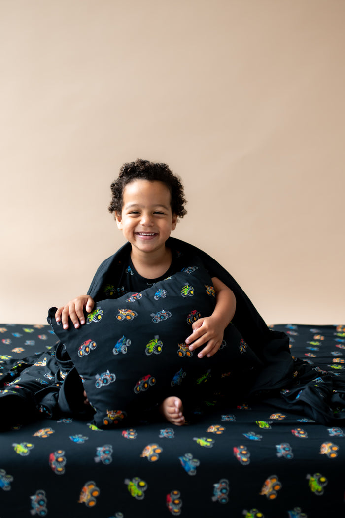 Boy holding black toddler pillowcase and wrapped in toddler blanket with Monster Truck print sitting on a similar print sheet