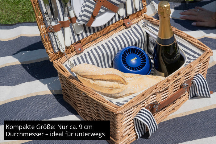 Ein Picknickkorb mit Brot, Sekt, Besteck und dem Unold Ventilator Breezy Bowl – einem kompakten Reiseventilator – auf einer gestreiften Decke. Der deutsche Text weist auf seine Handlichkeit und die perfekte Größe für unterwegs hin.
