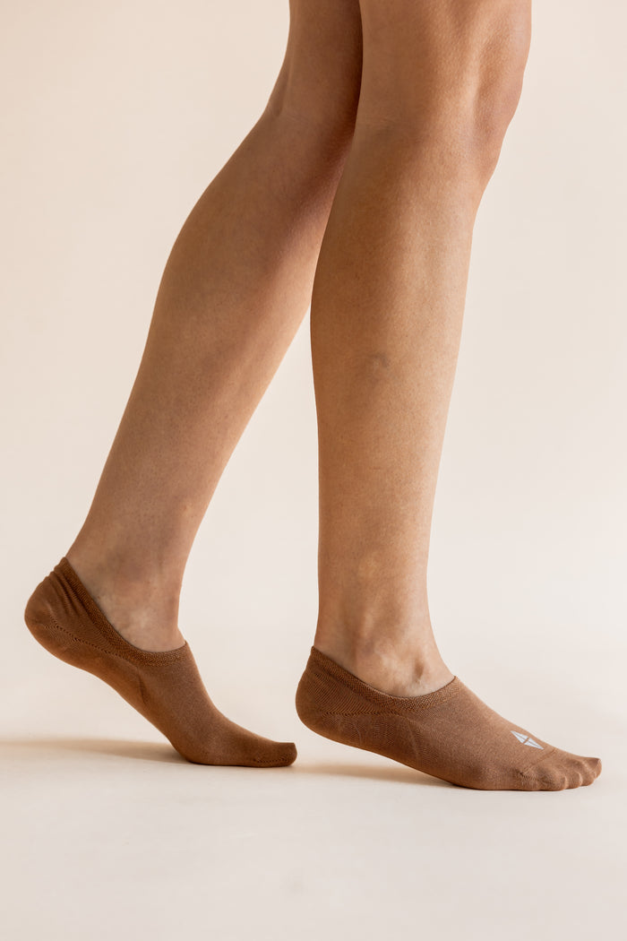 A close-up of a person's legs wearing brown no-show socks against a plain beige studio background.