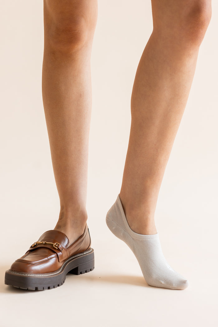 A close-up of a person's legs showing two different styles of low-cut socks: one leg wears a brown leather loafer with a chunky sole, while the other leg wears a light gray ankle sock.