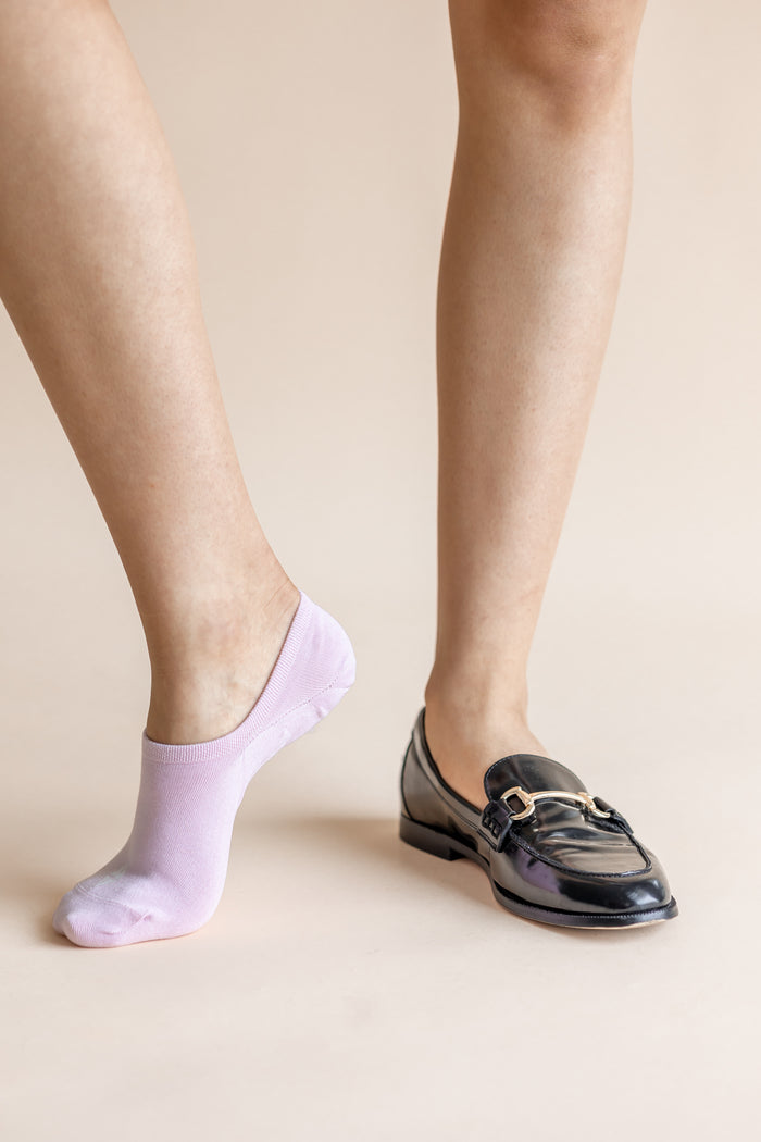 A person's legs shown from the calves down, wearing a light lavender no-show sock on one foot and a black patent leather loafer with a gold buckle on the other, against a solid beige background.