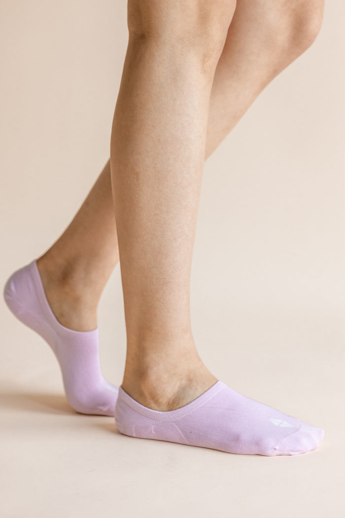 A close-up of a person's feet wearing light lavender low-cut no-show socks against a solid pale beige background.
