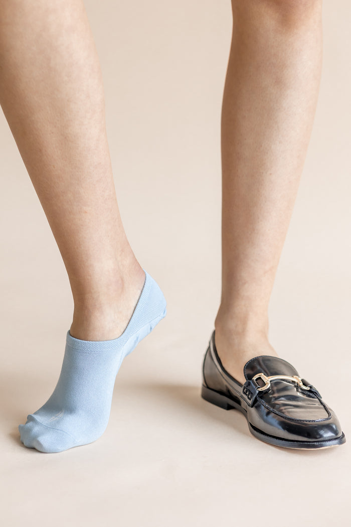 A close-up of a person's legs showing one ankle wearing a light blue no-show sock and the other wearing a black leather loafer with a gold buckle detail, set against a solid beige background.