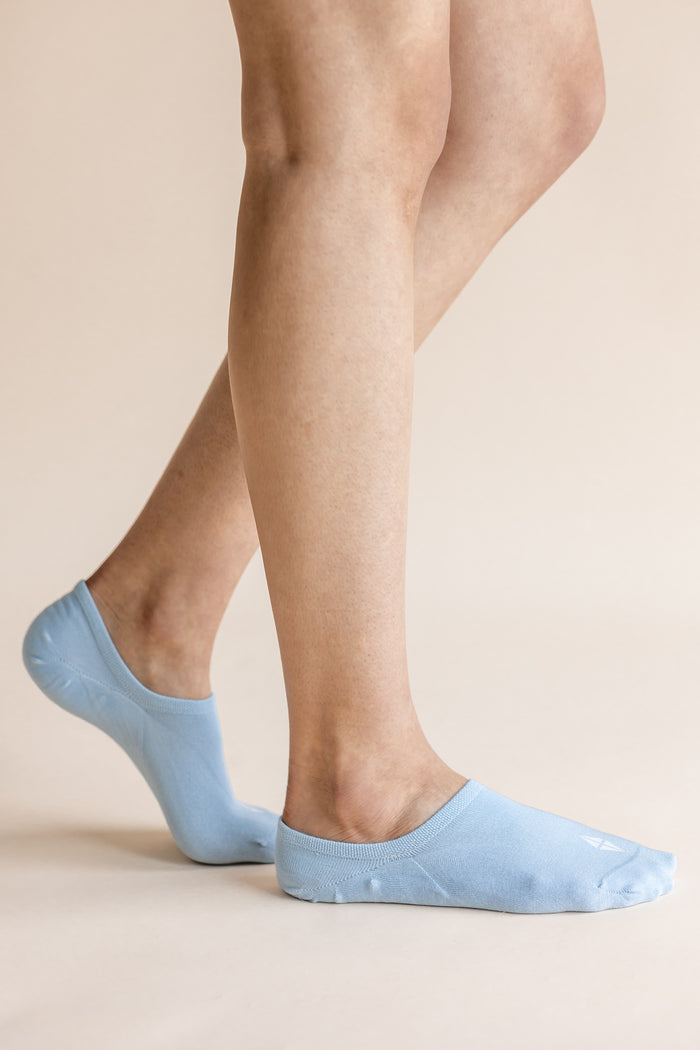A close-up of a person's legs wearing light blue low-cut no-show socks against a solid beige background.