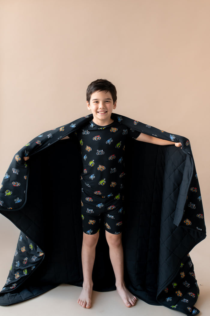 Boy holding black Youth Blanket in Monster Truck print wearing similar print short sleeve pajama set on a beige background