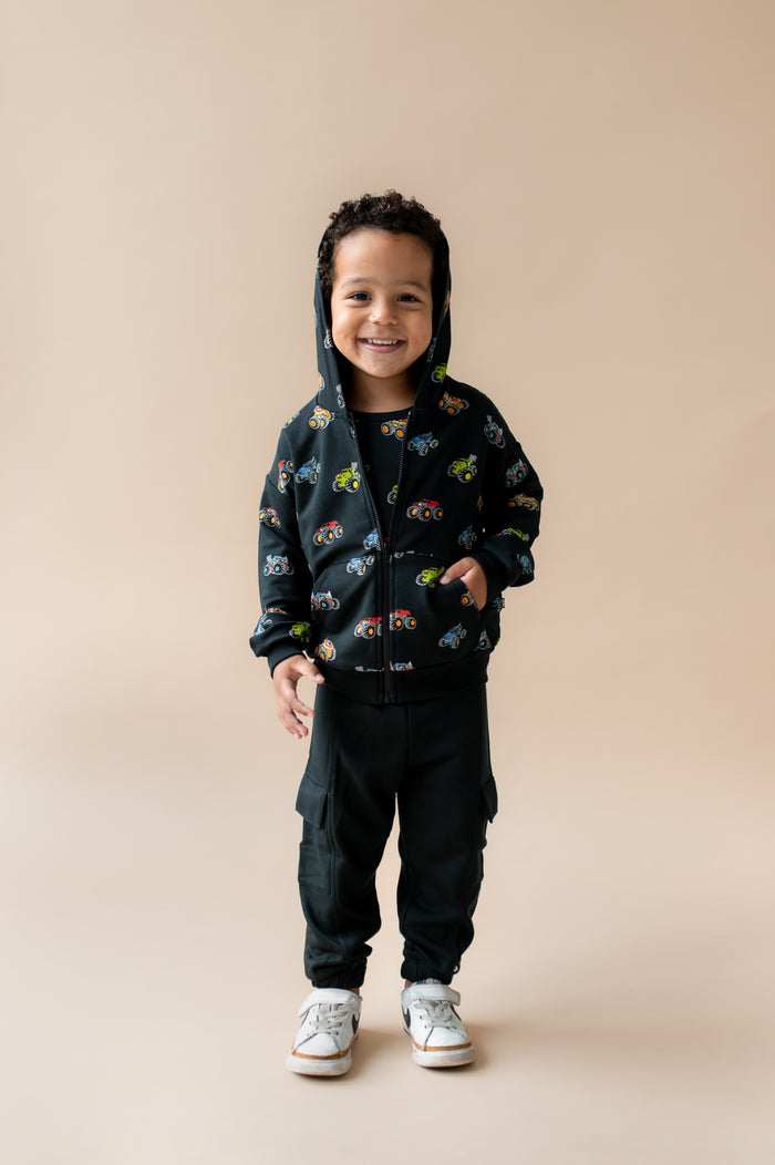 Boy standing wearing Black Zip-Up Hoodie in Monster Truck print over black pant and white shoes on a beige background