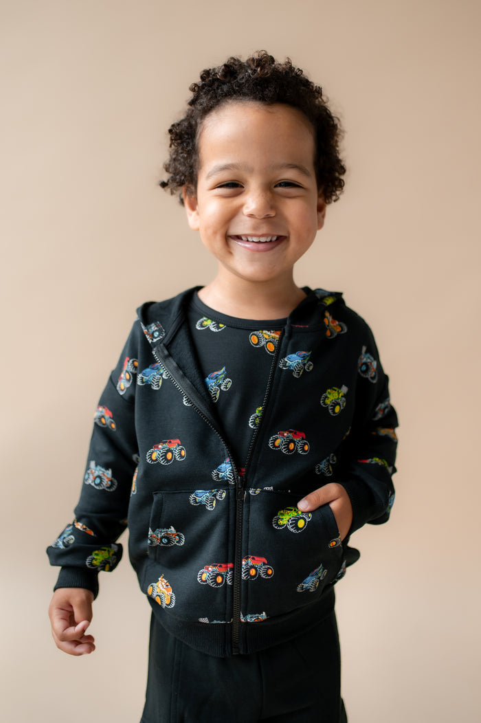 Boy smiling in Black Zip-Up Hoodie in Monster Truck print and black pant on a beige background