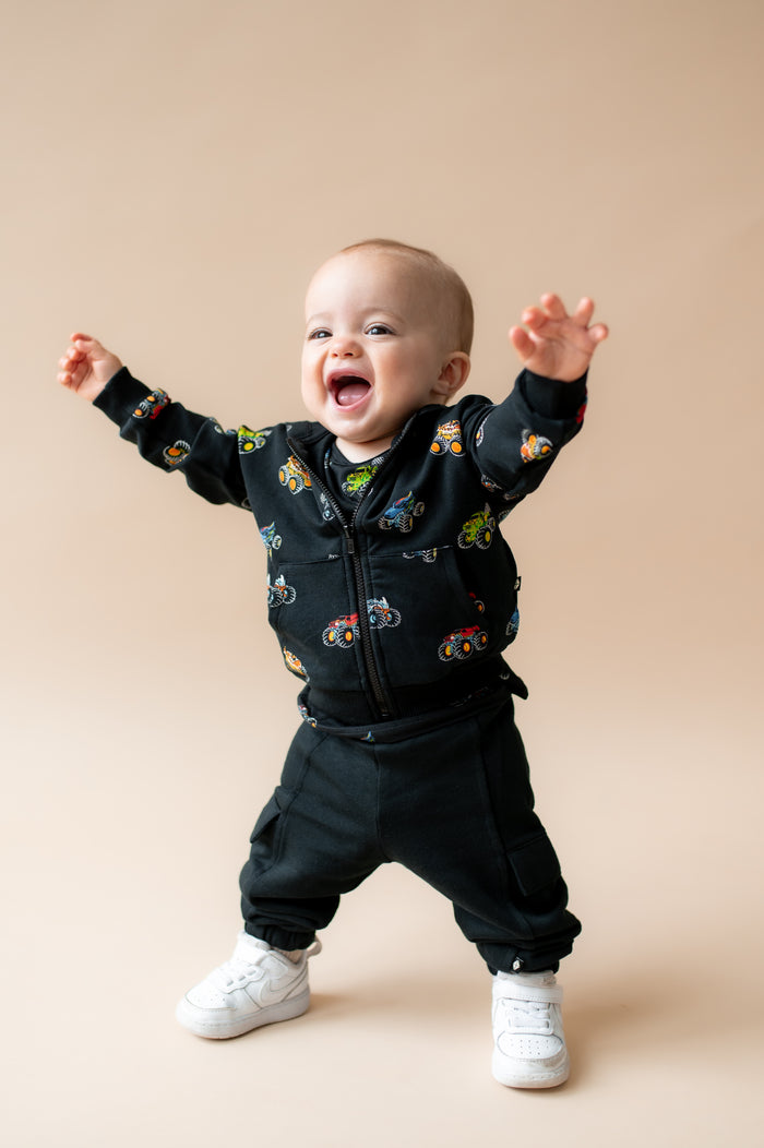 Toddler smiling in Black Zip-Up Hoodie in Monster Truck print over black pant and white shoes on a beige background