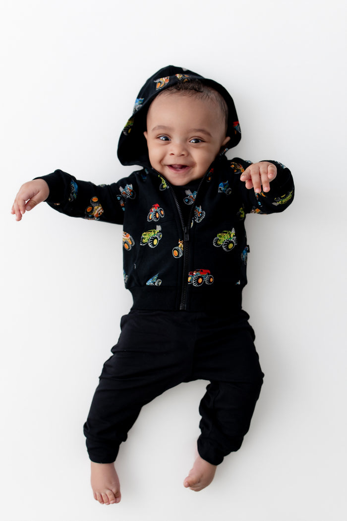 Baby wearing Black Zip-Up Hoodie in Monster Truck print and black pants on a white background