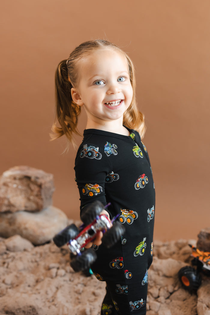 Girl holding toy truck smiling in black Zippered Romper in Monster Truck print on sand and rocks with toy trucks